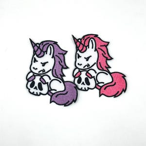 May include: Two embroidered patches featuring a unicorn design. One unicorn has purple hair and tail, the other has pink. Both unicorns are white with black horns and are holding a skull. The patches have a black border.