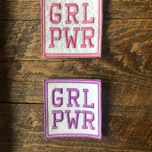May include: Two embroidered patches with the text "GRL PWR" in pink and purple lettering on a white background. The patches are surrounded by a pink and purple border.