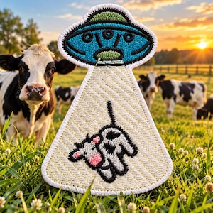 May include: An embroidered patch featuring a blue and green UFO hovering above a cream-colored beam of light. Inside the light beam, a small black and white cow with a pink nose is depicted in a floating position. The patch has a white border.