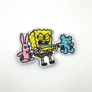 May include: Embroidered patch featuring SpongeBob SquarePants lifting a barbell with a pink bunny and a blue teddy bear attached. The patch has a white border and is on a white background. The cartoon characters are in bright colours.