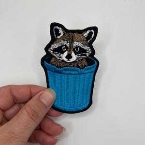 May include: Embroidered iron-on patch featuring a raccoon peeking out of a blue trash can.