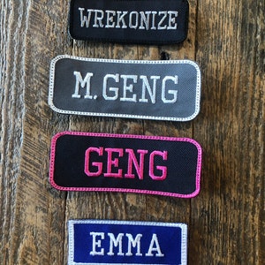 May include: Four embroidered patches with text on a wooden background. The patches are rectangular with rounded corners. The top patch is black with "WREKONIZE" in white. The second is gray with "M. GENG" in white. The third is black with "GENG" in pink. The bottom patch is blue with "EMMA" in white.