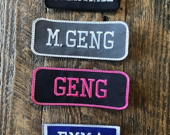 May include: Four embroidered patches with text on a wooden background. The patches are rectangular with rounded corners. The top patch is black with "WREKONIZE" in white. The second is gray with "M. GENG" in white. The third is black with "GENG" in pink. The bottom patch is blue with "EMMA" in white.