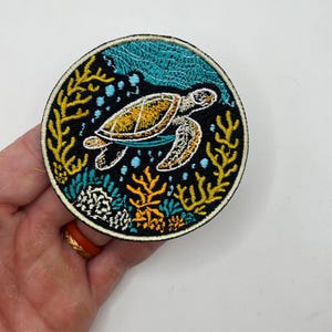 May include: Round embroidered patch depicting a sea turtle in an underwater scene. The patch has a black background, yellow coral, and a turquoise and yellow turtle. The patch is approximately 7.6 cm in diameter.