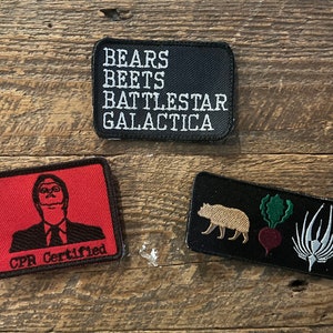 May include: Three rectangular embroidered patches. One black patch features the white text BEARS BEETS BATTLESTAR GALACTICA. A red patch shows a black illustration of a person with the text CPR Certified. A black patch displays a bear, a beet, and a symbol.