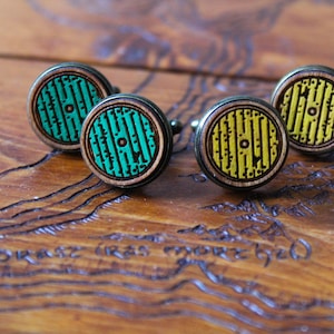 Può includere: Quattro gemelli in legno con un design circolare. Ogni gemello presenta un cerchio di colore diverso con un motivo di linee e un piccolo cerchio al centro. I colori sono verde, giallo, verde e giallo.