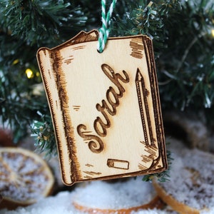 May include: A wooden ornament shaped like a closed book with a pencil and eraser drawn on it. The ornament is personalized with the name "Sarah" in cursive.