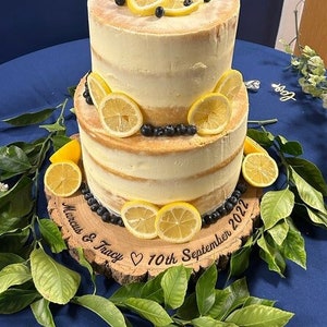 May include: A two-tiered lemon and blueberry wedding cake with a wooden cake stand. The cake stand is engraved with the text "Marcus & Tracy ♥ 10th September 2022".