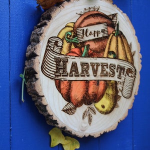 May include: A wooden round sign with a rustic bark edge, featuring a hand-drawn illustration of pumpkins, gourds, and leaves. The sign has the text "Happy Harvest" in a banner.
