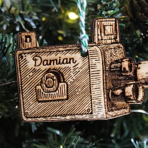 Może przedstawiać: Drewniana ozdoba choinkowa w kształcie vintage'owego aparatu fotograficznego z wygrawerowanym imieniem "Damian" na przodzie. Ozdoba ma brązowe wykończenie z drewna i jest ozdobiona misternymi detalami.