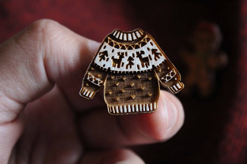 May include: A wooden Christmas ornament shaped like a brown sweater with white trim and a reindeer pattern.