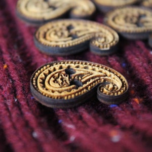 May include: A close-up of a set of wooden paisley-shaped buttons with a dark brown border. The buttons are on a burgundy fabric background.
