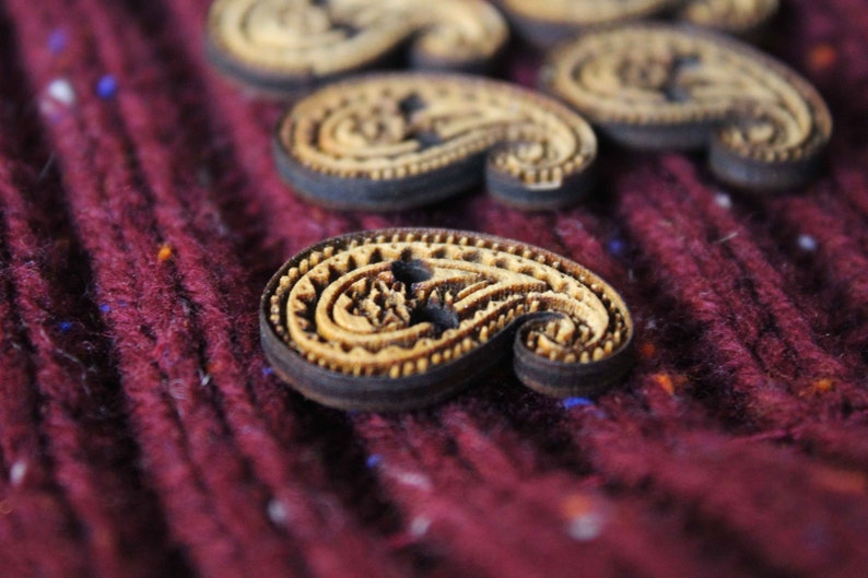 May include: A close-up of a set of wooden paisley-shaped buttons with a dark brown border. The buttons are on a burgundy fabric background.
