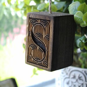 May include: A wooden block with the letter "S" carved into it. The letter is surrounded by a decorative pattern of lines and curves. The block is hanging from a string.