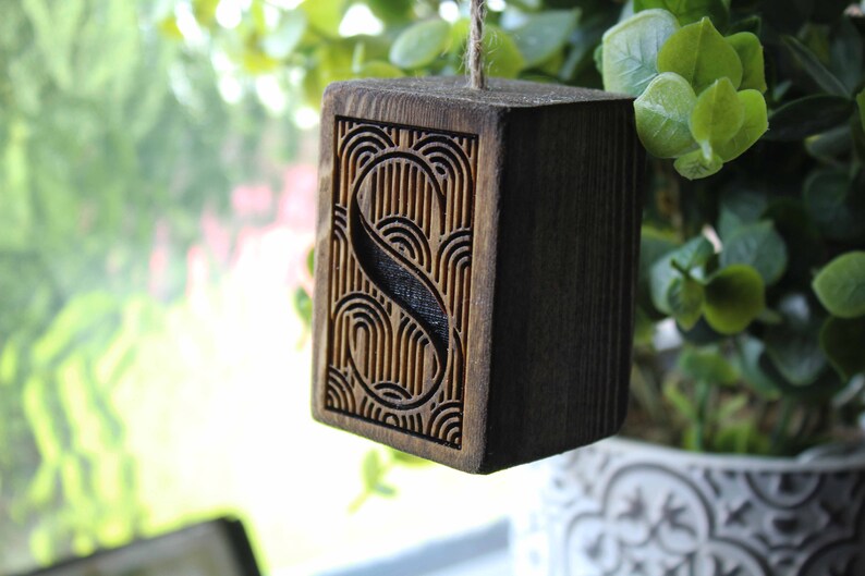 May include: A wooden block with the letter "S" carved into it. The letter is surrounded by a decorative pattern of lines and curves. The block is hanging from a string.