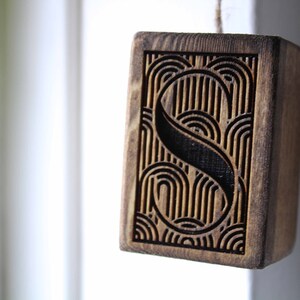 May include: A wooden block with a geometric pattern carved into it. The pattern features a stylised letter "S" surrounded by curved lines.