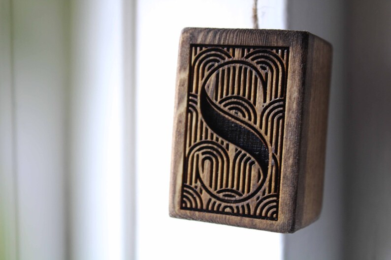 May include: A wooden block with a geometric pattern carved into it. The pattern features a stylised letter "S" surrounded by curved lines.