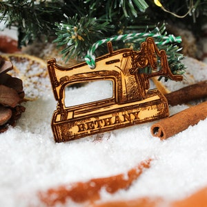 May include: A wooden Christmas bauble shaped like a vintage sewing machine with the name "Bethany" engraved on the base. The bauble is surrounded by pine branches, cinnamon sticks, and faux snow.
