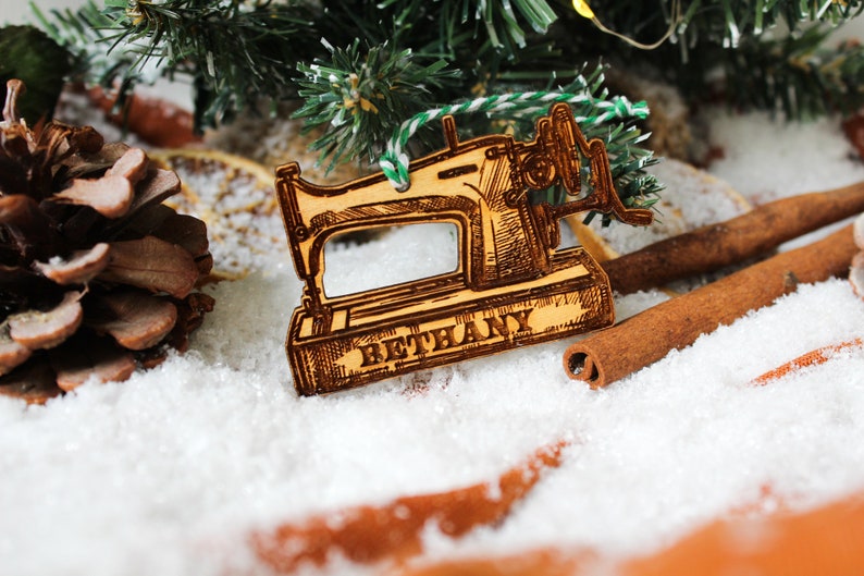 May include: A wooden Christmas bauble shaped like a vintage sewing machine with the name "Bethany" engraved on the base. The bauble is surrounded by pine branches, cinnamon sticks, and faux snow.