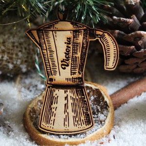 May include: A wooden Christmas bauble shaped like a coffee pot with the name "Victoria" engraved on it. The bauble is sitting on a dried orange slice and surrounded by pine cones and cinnamon sticks.