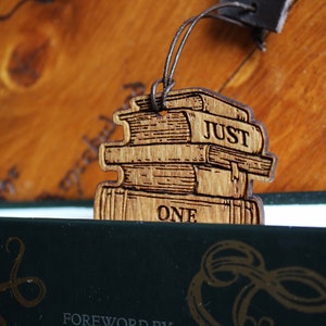 May include: A wooden bookmark shaped like a stack of books with the words "Just One" engraved on the bottom book. The bookmark is hanging from a brown leather cord.