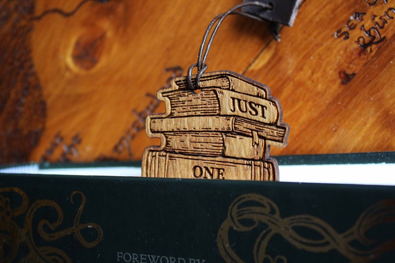 May include: A wooden bookmark shaped like a stack of books with the words "Just One" engraved on the bottom book. The bookmark is hanging from a brown leather cord.