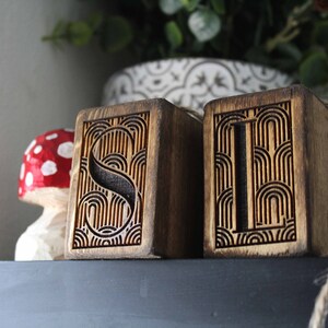 May include: Two wooden blocks with the letters "S" and "I" carved into them. The letters are surrounded by a decorative pattern of curved lines. The blocks are stained a dark brown colour.