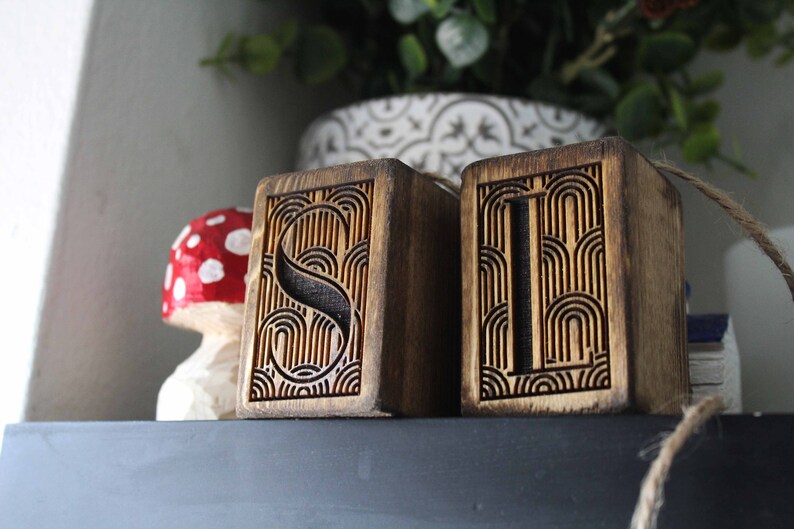 May include: Two wooden blocks with the letters "S" and "I" carved into them. The letters are surrounded by a decorative pattern of curved lines. The blocks are stained a dark brown colour.