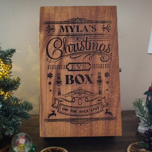 May include: A wooden box with the text "Myla's Christmas Eve Box On The Nice List" engraved on the front. The box is brown and has a rustic look.