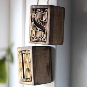 May include: Two wooden blocks with the letters "S" and "I" carved into them. The blocks are hanging from a string and have a geometric pattern carved into the surface.