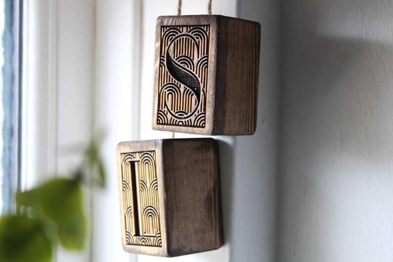 May include: Two wooden blocks with the letters "S" and "I" carved into them. The blocks are hanging from a string and have a geometric pattern carved into the surface.
