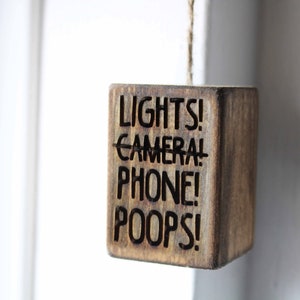 Peut inclure: Une pancarte en bois avec le texte "LIGHTS! CAMERA! PHONE! POOPS!" suspendue à une ficelle.