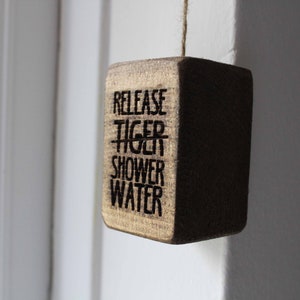 Pode incluir: Uma placa de madeira com o texto "Release Tiger Shower Water" pendurada em uma porta branca.
