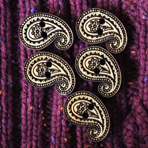 May include: Five wooden paisley-shaped buttons with black and white designs. The buttons are arranged in a circle on a dark purple fabric background.