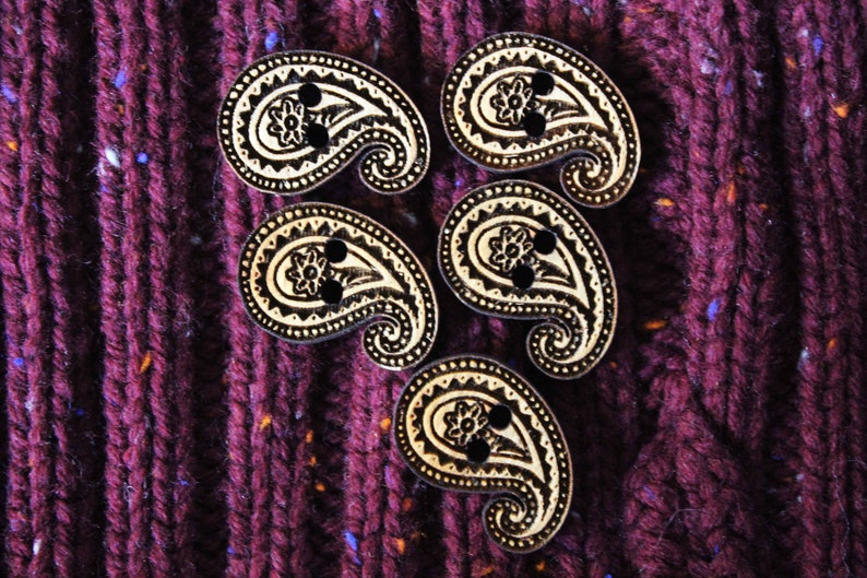 May include: Five wooden paisley-shaped buttons with black and white designs. The buttons are arranged in a circle on a dark purple fabric background.