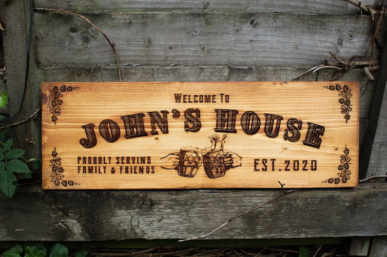 May include: A wooden sign with the text "Welcome to John's House" and "Proudly Serving Family & Friends" with two beer mugs clinking together. The sign also includes the text "Est. 2020".