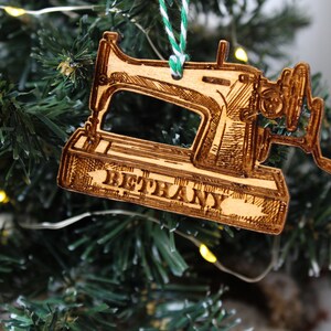 May include: A wooden Christmas bauble shaped like a vintage sewing machine with the name "Bethany" engraved on the base. The bauble is hanging on a green Christmas tree.