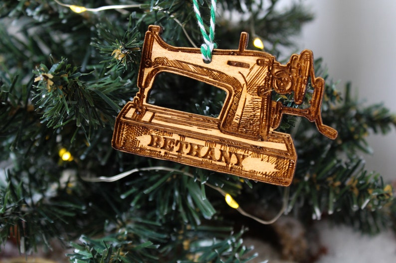 May include: A wooden Christmas bauble shaped like a vintage sewing machine with the name "Bethany" engraved on the base. The bauble is hanging on a green Christmas tree.