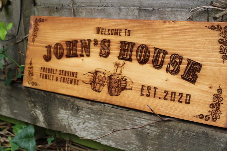 May include: Wooden sign with the text "Welcome to John's House, Probably Serving Family & Friends, Est. 2020". The sign is decorated with a vine and leaf design and two beer mugs.