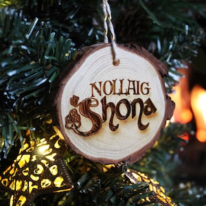 May include: A wooden Christmas bauble with the text "Nollaig Shona" in a decorative font. The bauble is hanging on a green Christmas tree.
