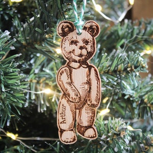 May include: A wooden Christmas ornament shaped like a teddy bear with the name "Arthur" engraved on it. The ornament is hanging on a green Christmas tree.