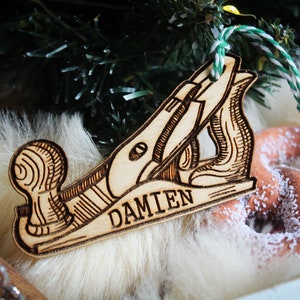 May include: Wooden ornament shaped like a hand plane, with the name "DAMIEN" engraved. The ornament is light brown with dark brown etched details and a green and white string for hanging. The background includes a Christmas tree branch and a pretzel.