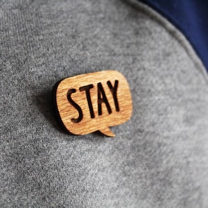 Pode incluir: Um alfinete de madeira em forma de balão de fala com a palavra "STAY" gravada nele. O alfinete está preso a uma camisa cinza.