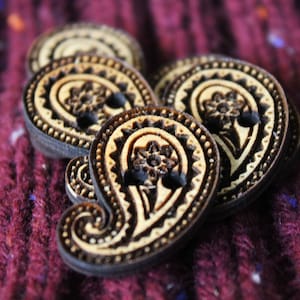 May include: Four wooden buttons with intricate paisley designs in brown and cream colours. The buttons are arranged in a pile on a burgundy knitted fabric.