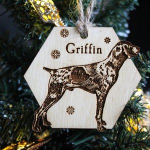 May include: A wooden hexagonal Christmas ornament with a laser-engraved image of a dog and the name "Griffin". The ornament has a snowflake design and is hanging on a Christmas tree.