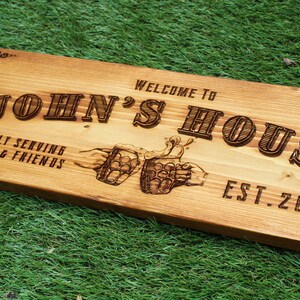 May include: Wooden sign with the text "Welcome to John's House, Proudly Serving Family & Friends, Est. 2020". The sign features a design of two hands clinking beer mugs.