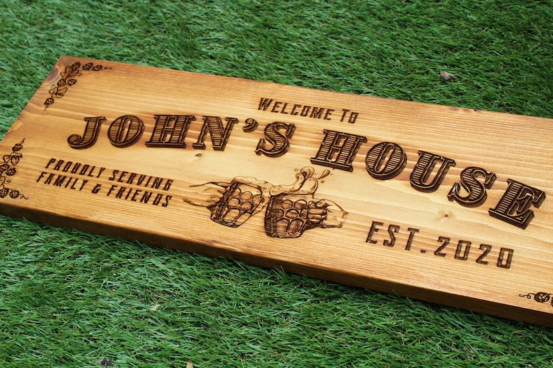 May include: Wooden sign with the text "Welcome to John's House, Proudly Serving Family & Friends, Est. 2020". The sign features a design of two hands clinking beer mugs.