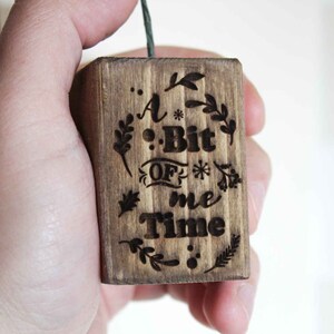 Könnte beinhalten: Ein Holzblock-Anhänger mit dem Text "A Bit Of Me Time" und eingravierten Blumenmustern.