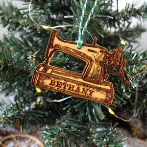 May include: A wooden Christmas bauble shaped like a vintage sewing machine with the name "Bethany" engraved on the base. The bauble is hanging on a green Christmas tree.