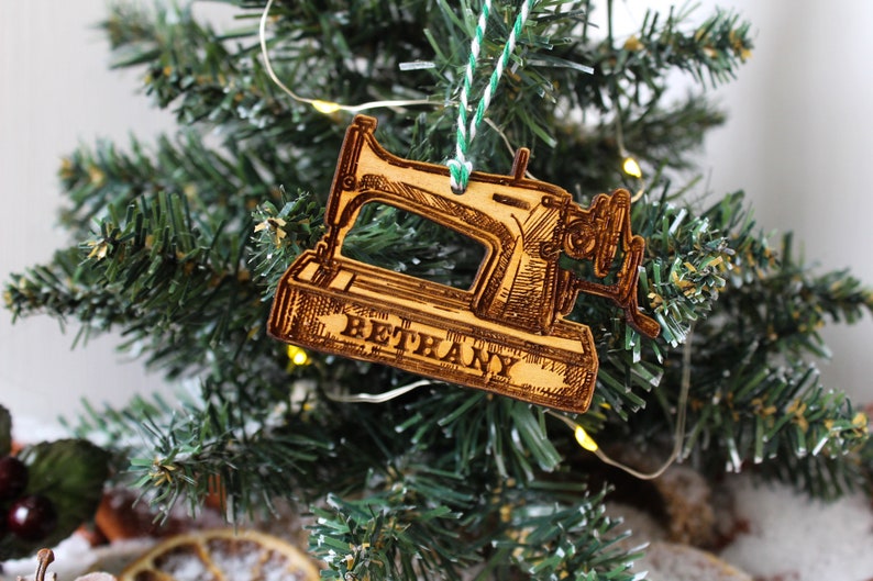 May include: A wooden Christmas bauble shaped like a vintage sewing machine with the name "Bethany" engraved on the base. The bauble is hanging on a green Christmas tree.